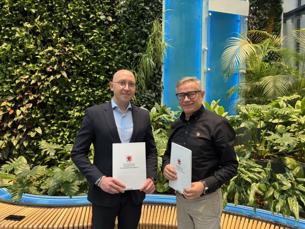 Wicemarszałek Województwa Zachodniopomorskiego Jakub Kowalik i dyrektor Ogrodów Przelewice Zygmunt Siarkiewicz stoją na tle ściany pokrytej roślinnością w siedzibie Urzędu marszałkowskiego Województwa Zachodniopomorskiego, trzymając teczki z herbem Województwa Zachodniopomorskiego